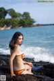 A woman in an orange bikini sitting on a rock by the ocean.